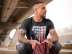 Man wearing Fight Fight Fight Bold Logo Tee with patriotic design, sitting under an urban bridge.