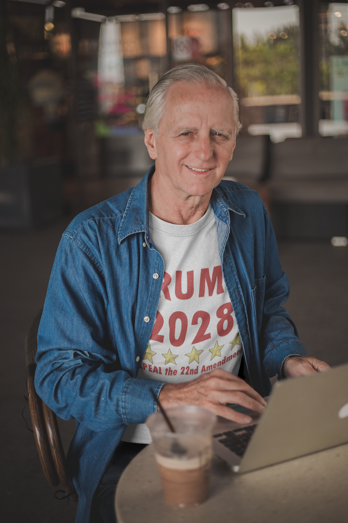Man wearing a Trump 2028 t-shirt advocating for the 2028 Repeal 22nd Amendment Trump while using a laptop.