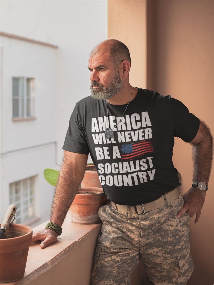 Man wearing 'America Will Never Be A Socialist Country' t-shirt, standing on balcony with potted plants, confident expression