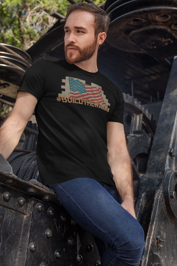 Man wearing a black t-shirt with 'Build The Wall Bold Statement Design' in front of industrial background