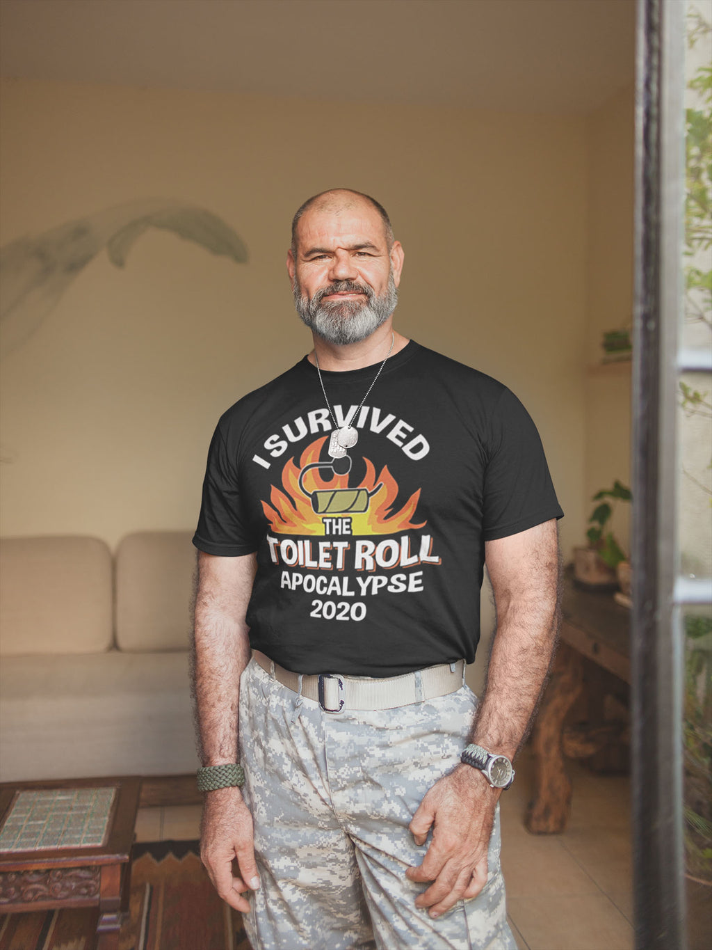Man wearing 'I Survived The Toilet Roll Apocalypse 2020' t-shirt, showcasing a humorous design, in a cozy living room.