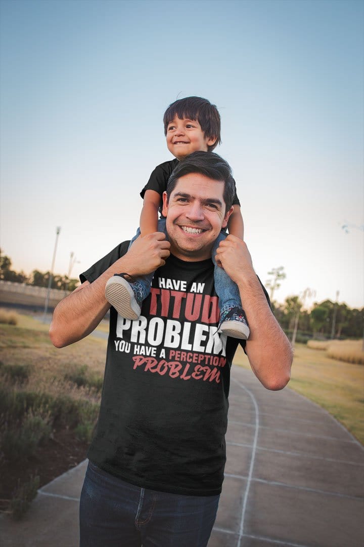 Man wearing Attitude Problem Funny Graphic Tee carrying child on shoulders outdoors.