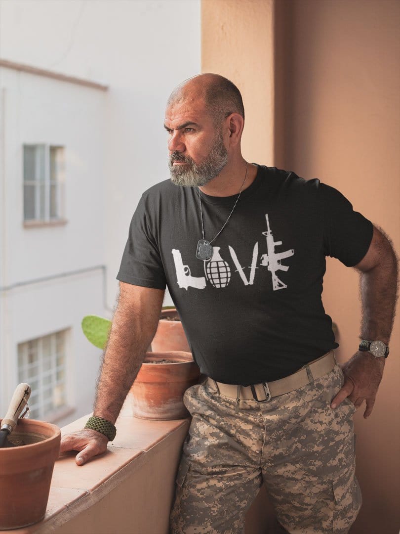 Man wearing 'Love As Pistol Grenade Knives And AR-15' graphic t-shirt, standing on a balcony in casual camo pants.