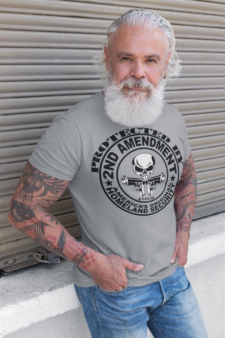 Man wearing 'Protected by 2nd Amendment Homeland Security' t-shirt with tattoos, standing outdoors.