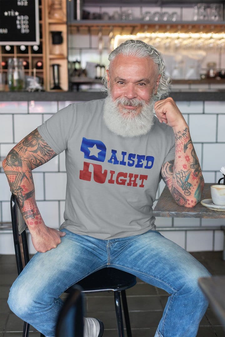 Man wearing 'Raised Right Inspirational Graphic Design' shirt, sitting in a cafe.
