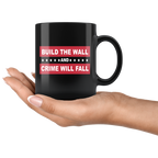 Hand holding Border Security Equals Safer Land black mug with 'Build The Wall And Crime Will Fall' text.